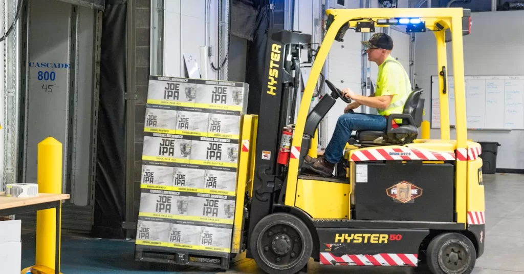 A worker drives a Hyster forklift moving Joe IPA boxes in a warehouse.