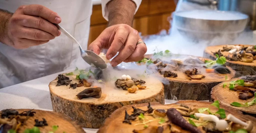 A chef artfully plating a gourmet dish with mushrooms and greens on wood slices.