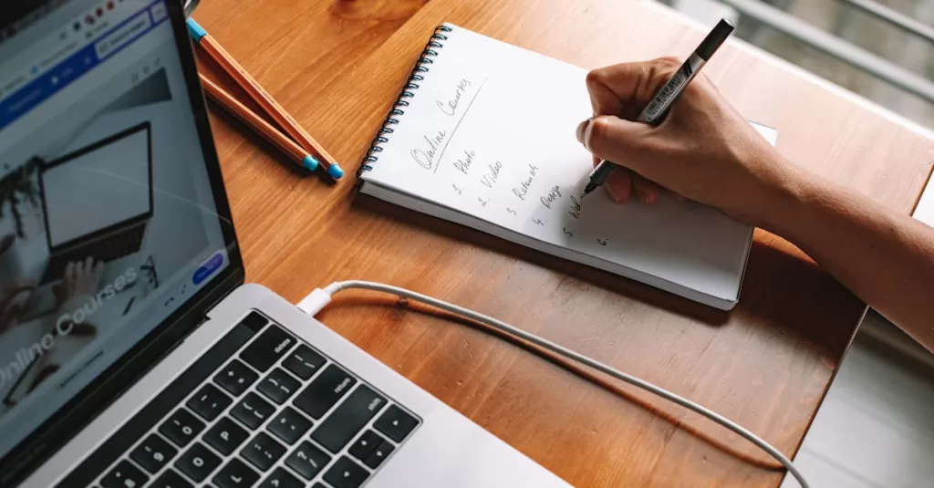 A person writing notes while researching online courses on a laptop in a home setting.