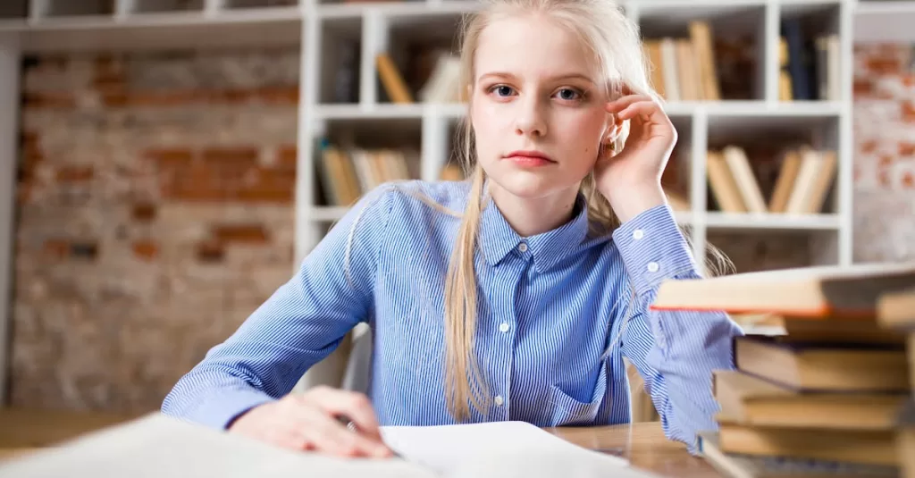 A young woman with blond hair studying at a table piled with books.