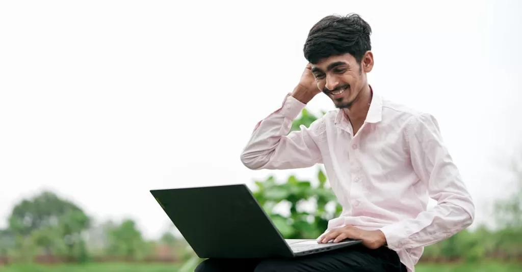 Young man in a pink shirt smiling while using a laptop outdoors in a lush green park setting.
