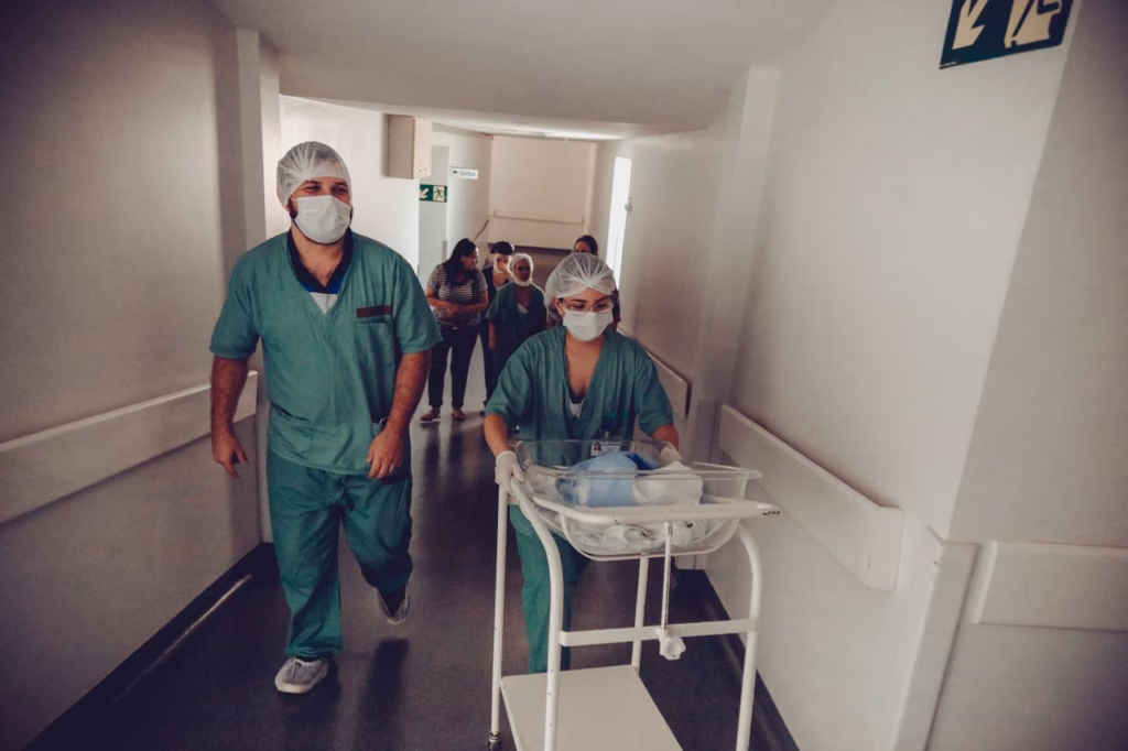 Anonymous male gynecologist in uniform and female colleague in sterile mask carrying medical trolley with newborn baby in corridor in clinic behind group of people